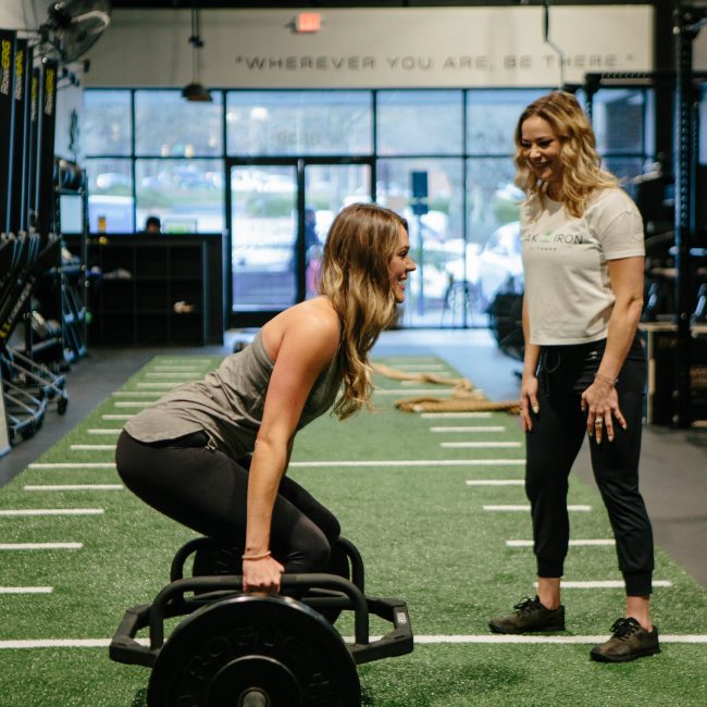 With the camera pointed towards the front of the gym, Trainer Kayla Scott coaches a personal training client in the deadlift.