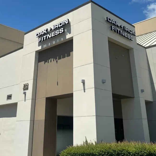 Exterior view of Oak and Iron Fitness gym building in Cary, North Carolina, showing a modern brick-and-glass structure with the gym’s signage visible above the entrance. Locations